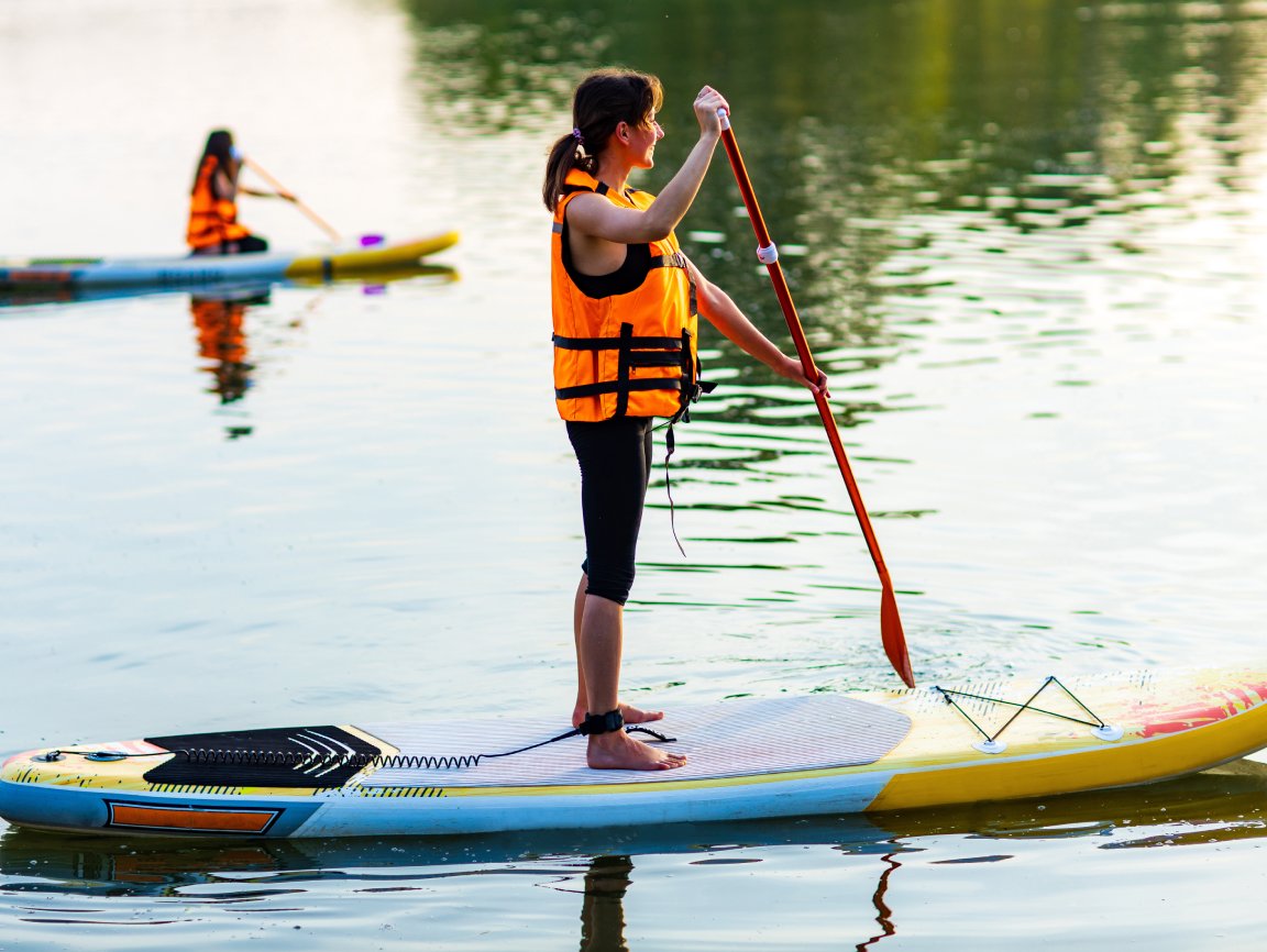 A woman wearing a yellow life jacket stands on a paddleboard. She's about to dip her paddle into the water.