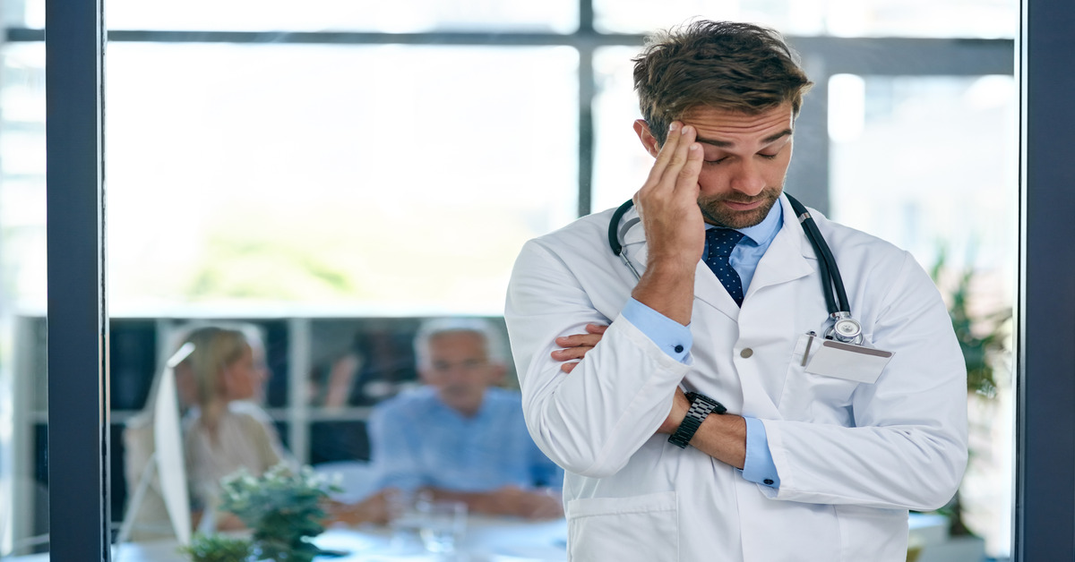 Physician in a white lab coat standing in a clinic has his hand on his temple with nursing staff behind him.