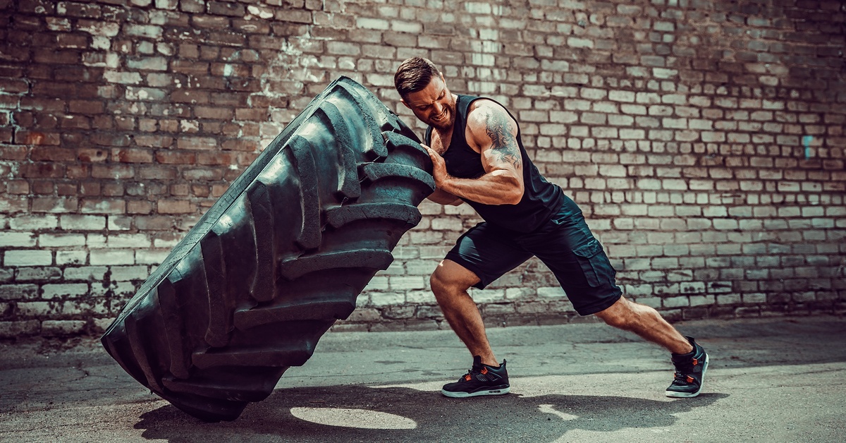 An athletic person with an arm tattoo using their body to flip a massive black tire while outside a brick building.