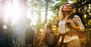 A group hikes through a sunlit forest as beams of light stream through the trees. A woman looks up and smiles in amazement.