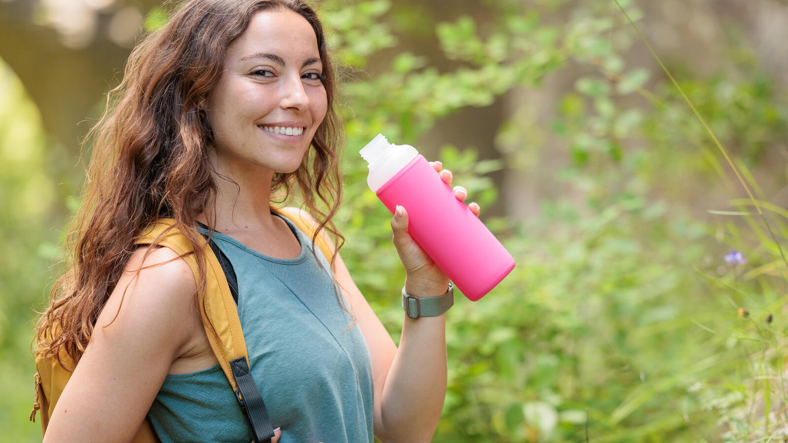 A smiling brunette woman wearing a blue camisole and yellow backpack smiles and holds a pink water bottle on a hiking trail.