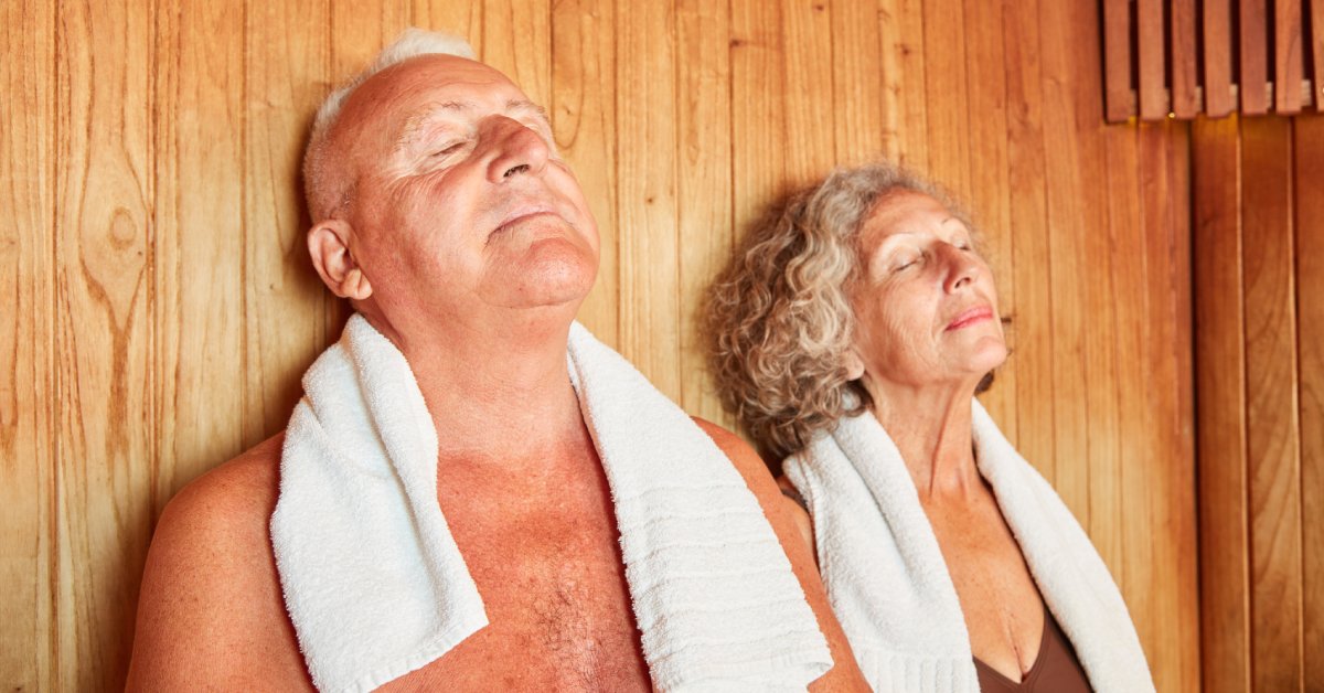 Two seniors, one male and one female, relax in a wooden sauna with their eyes closed and towels around their necks.