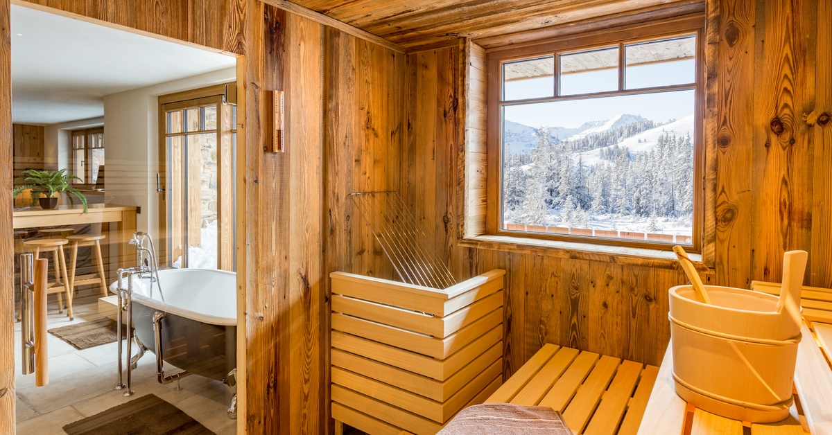 A sauna in a separate room of a home next to a seating area with a luxurious basin. A window reveals an alpine scenery.