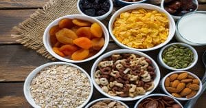 Snack for Post-Workout Recovery, Several white bowls holding dried fruit, seeds, cereal, oats, and nuts sitting on a wooden table with burlap fabric nearby.