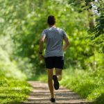A man in shorts and a T-shirt jogs alone down a narrow trail through a green wooded area on a sunny day.