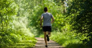 A man in shorts and a T-shirt jogs alone down a narrow trail through a green wooded area on a sunny day.