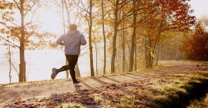 A man in a gray hoodie is shown running on a tree-lined trail early in the morning. The trees have mostly shed their autumn leaves.