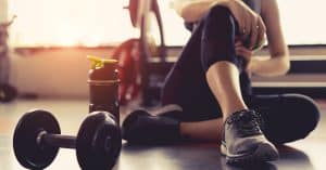 A woman sitting on the ground, her right arm resting on her right knee. There is a water bottle and a dumbbell next to her.