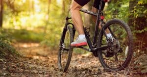 The lower half of a man as he bikes on a trail. He's wearing biker shorts and shoes with bright pink soles.