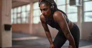 A woman wearing a cropped workout shirt, long pants, and headphones rests her body while taking a workout break.