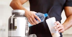 A muscular person in a black shirt pouring water into a plastic cup that already contains a pale protein powder.