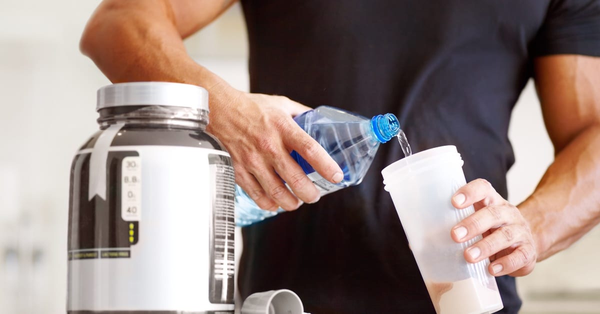 A muscular person in a black shirt pouring water into a plastic cup that already contains a pale protein powder.