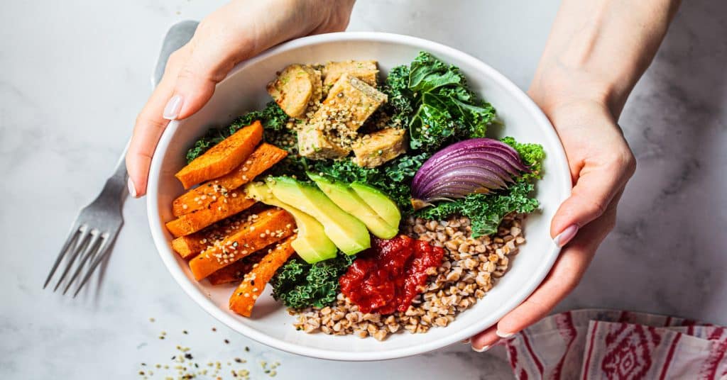 Someone holding a white bowl full of colorful foods, including sliced avocado, tofu, kale, seeds, and other healthy ingredients.