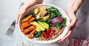 Someone holding a white bowl full of colorful foods, including sliced avocado, tofu, kale, seeds, and other healthy ingredients.