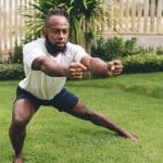A man in a T-shirt and shorts exercising without equipment in an outdoor area full of lush green grass.