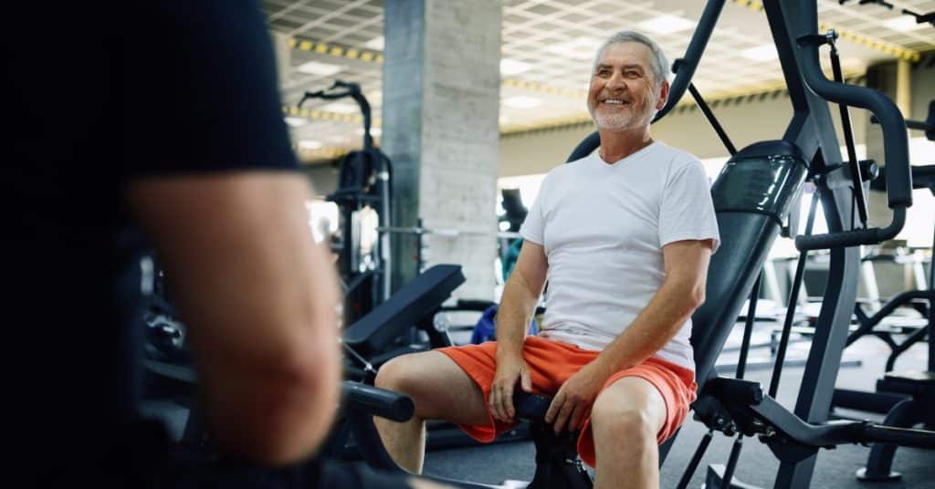 An elderly man sitting at a workout station, talking to another person. He is wearing bright orange shorts.