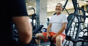 An elderly man sitting at a workout station, talking to another person. He is wearing bright orange shorts.