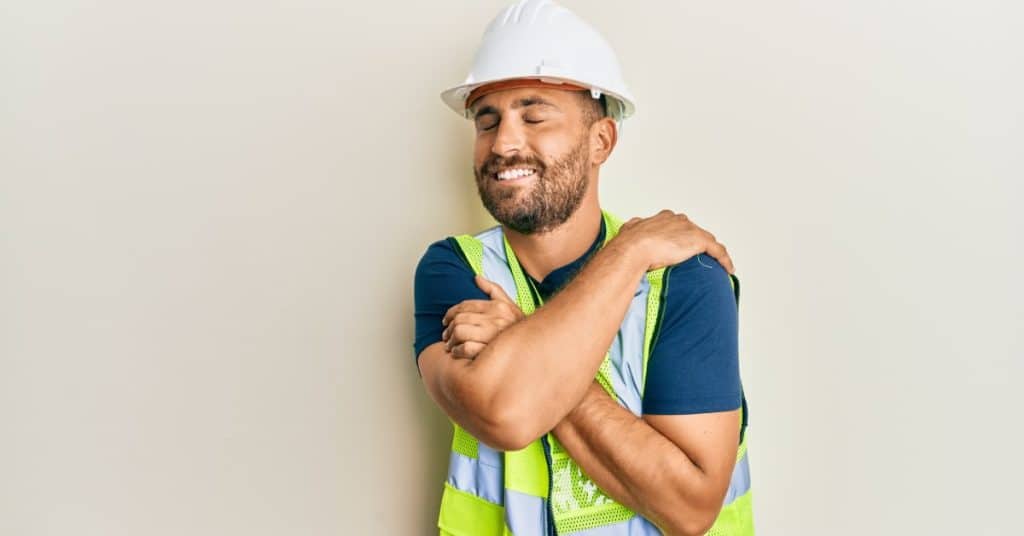 An adult male wearing a white hard hat and a reflective safety vest closes his eyes and smiles as he hugs himself.