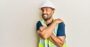An adult male wearing a white hard hat and a reflective safety vest closes his eyes and smiles as he hugs himself.