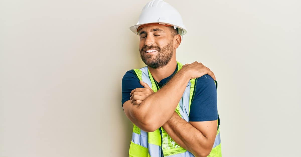 An adult male wearing a white hard hat and a reflective safety vest closes his eyes and smiles as he hugs himself.