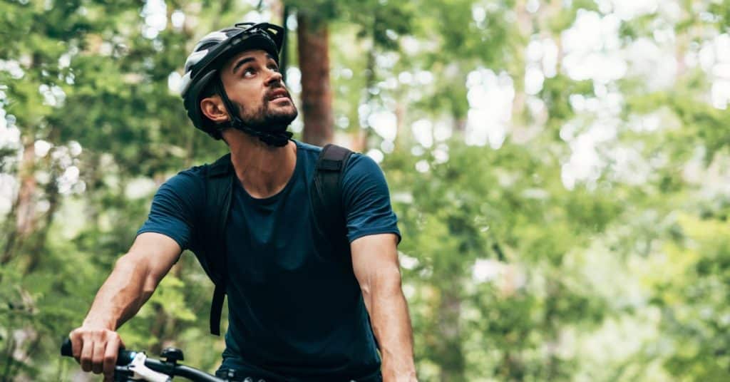 A man wearing a helmet while riding a bike outdoors looking upwards in a forest setting with many trees surrounding him.