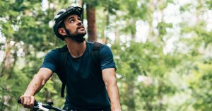 A man wearing a helmet while riding a bike outdoors looking upwards in a forest setting with many trees surrounding him.
