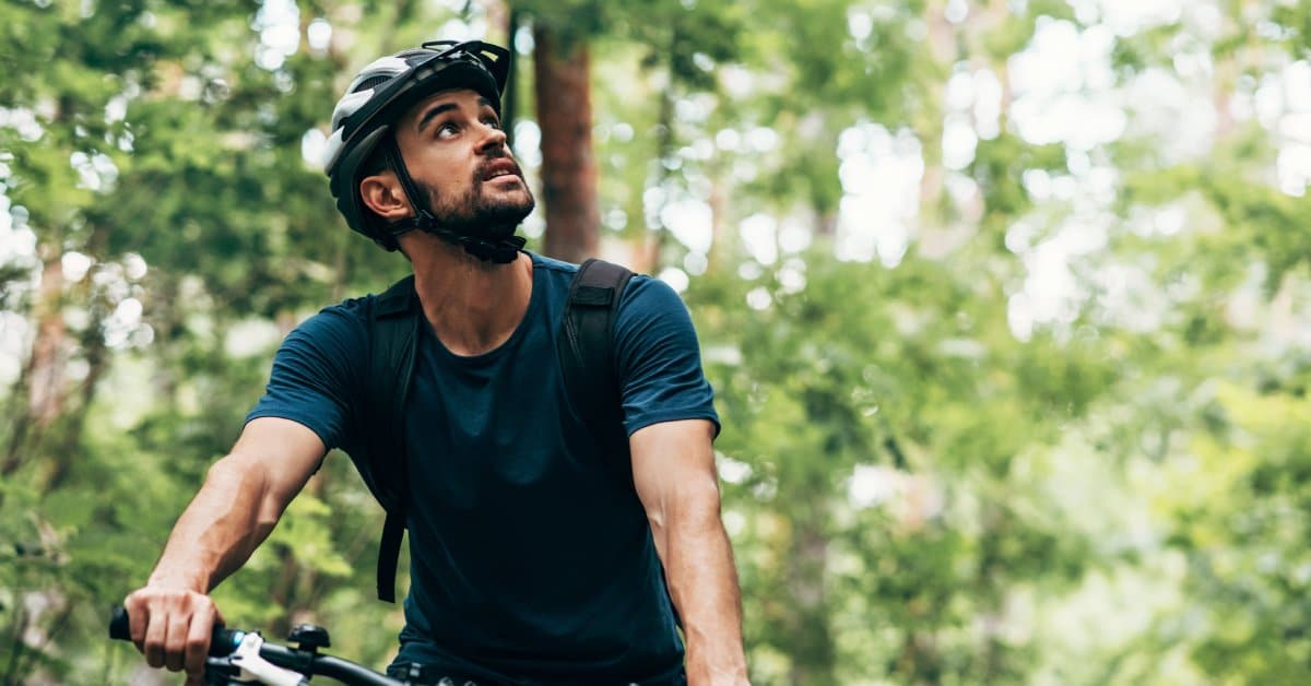 A man wearing a helmet while riding a bike outdoors looking upwards in a forest setting with many trees surrounding him.