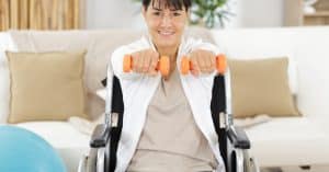 A woman wearing athletic clothing, sitting in a wheelchair in her living room, holding small orange dumbbells in front of her.