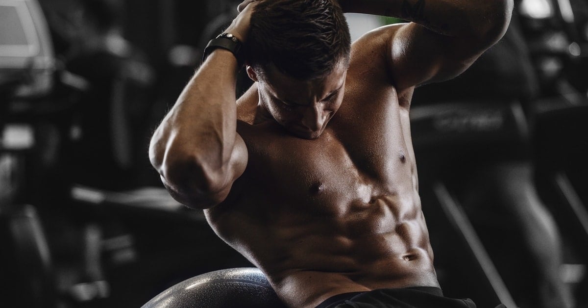 An athletic man doing crunches while balancing on an exercise ball in a gym. Other equipment sits in the background.