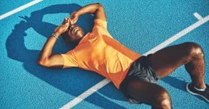 A young male athlete lies on his back on a blue track. He places his hands over his eyes, appearing fatigued.