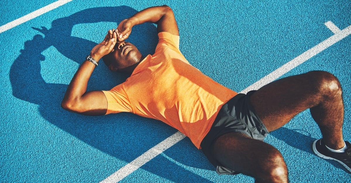 A young male athlete lies on his back on a blue track. He places his hands over his eyes, appearing fatigued.
