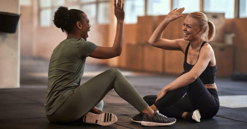 Two women wearing matching exercise sets sit on a black gym floor, laughing and high-fiving each other.