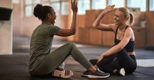 Two women wearing matching exercise sets sit on a black gym floor, laughing and high-fiving each other.