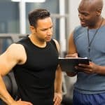 A personal trainer in athletic wear holding a clipboard and turning to speak to a client. Both are in a workout area.