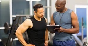 A personal trainer in athletic wear holding a clipboard and turning to speak to a client. Both are in a workout area.