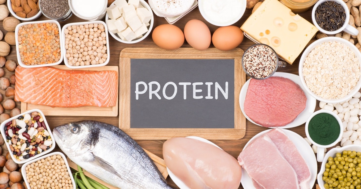 A variety of protein food sources on a wooden board. There is a chalkboard sign in the middle that reads "Protein."