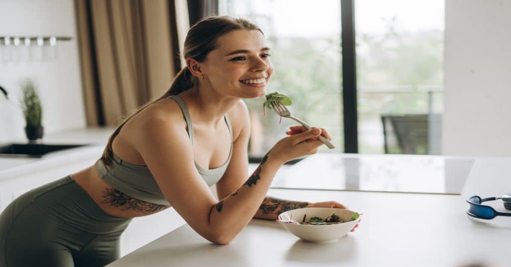 A young woman in workout gear leans against a counter with a bowl of salad in front of her. She holds a fork to her mouth.