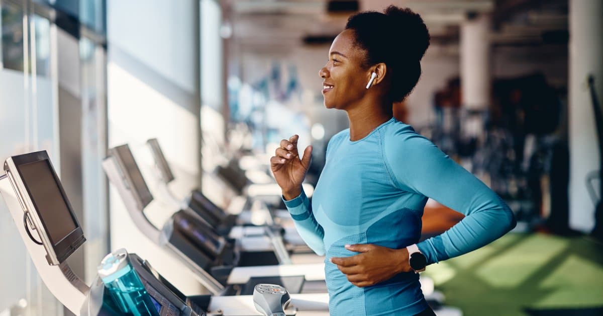 A Black woman is happily running on a treadmill with an AirPod in one ear. She's wearing bright colored athletic gear.