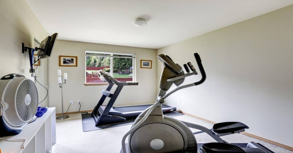 The interior of a small home gym with a treadmill, an elliptical, shelves, a fan, and a TV mounted on the wall.