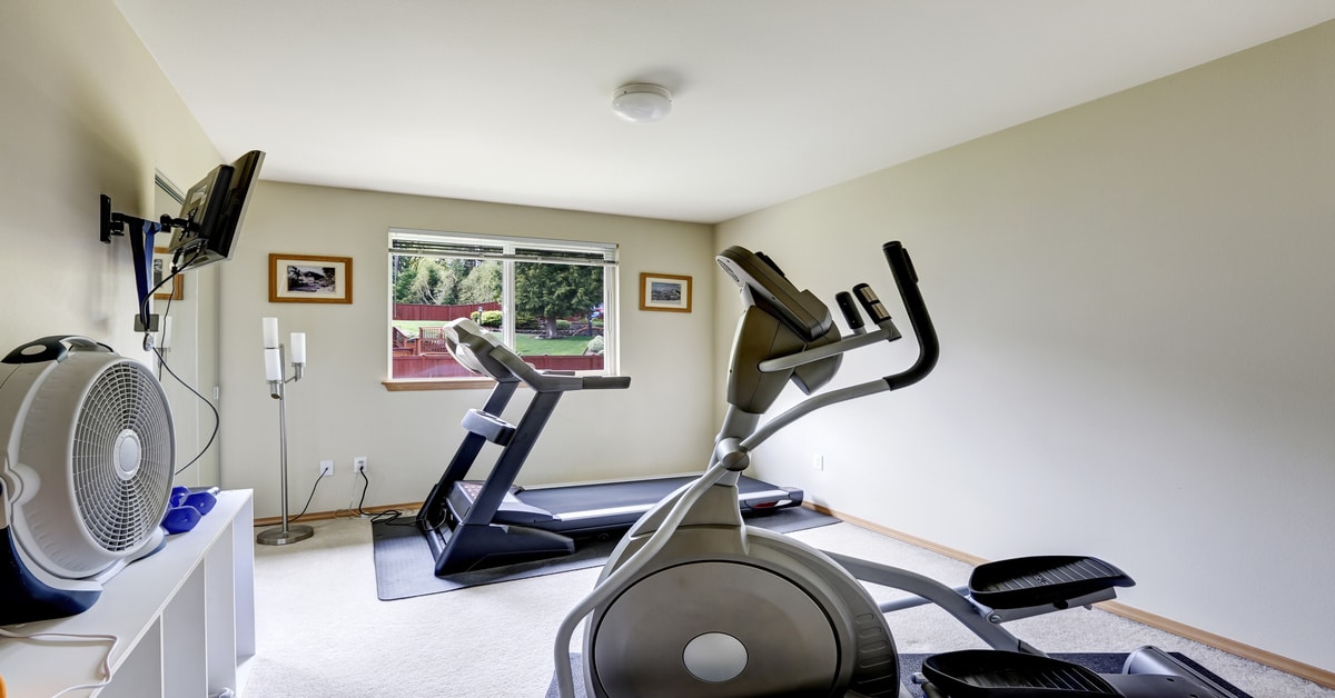 The interior of a small home gym with a treadmill, an elliptical, shelves, a fan, and a TV mounted on the wall.