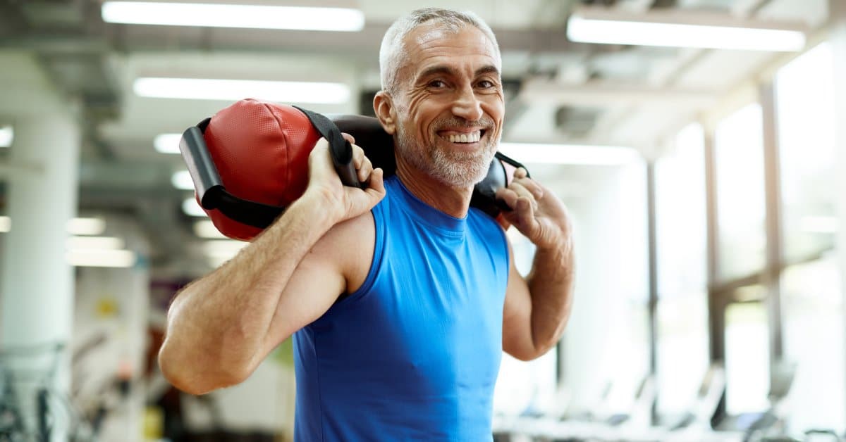 A mature man is smiling at the camera as he works out and lifts a weighted bag in a private gym setting.
