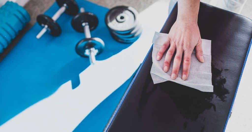 A hand wiping a disinfecting wipe across the surface of a cushioned bench. A yoga mat with weights is spread out below.