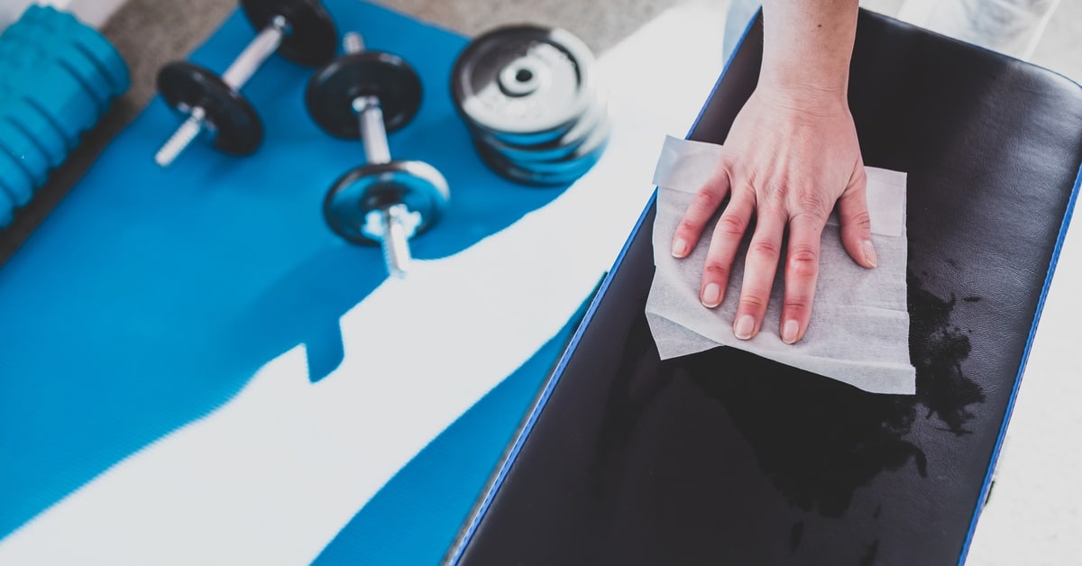 A hand wiping a disinfecting wipe across the surface of a cushioned bench. A yoga mat with weights is spread out below.