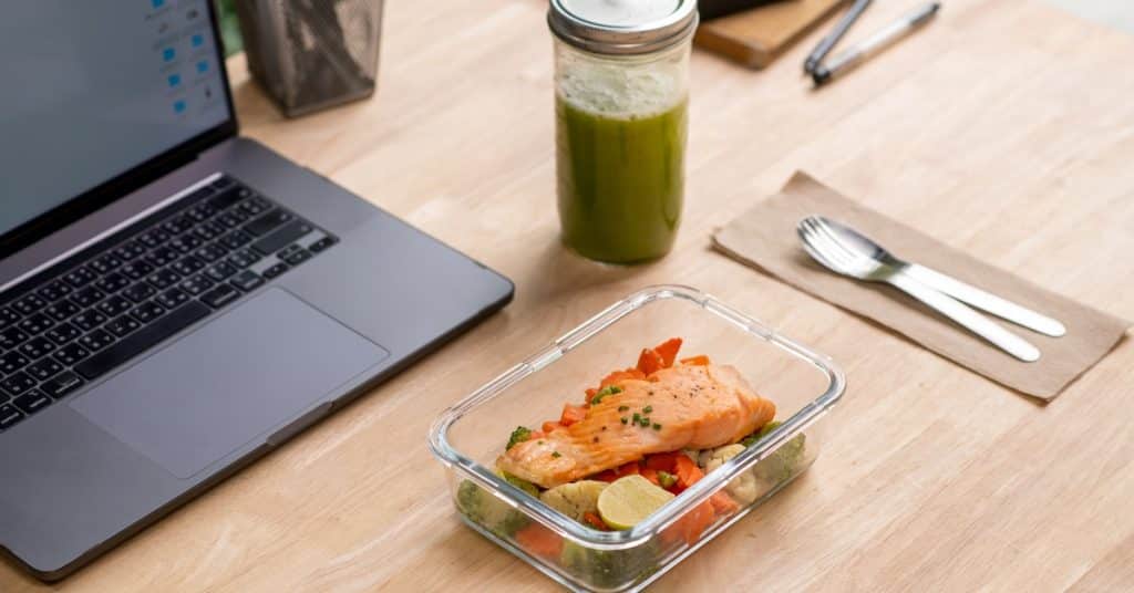 A glass container on a wooden desk contains salmon and vegetables. Silverware, a glass cup, and a laptop surround the food.
