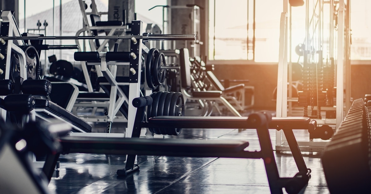 The interior of a gym with windows and various exercise equipment such as black benches, weights, and lat pulldown machines.
