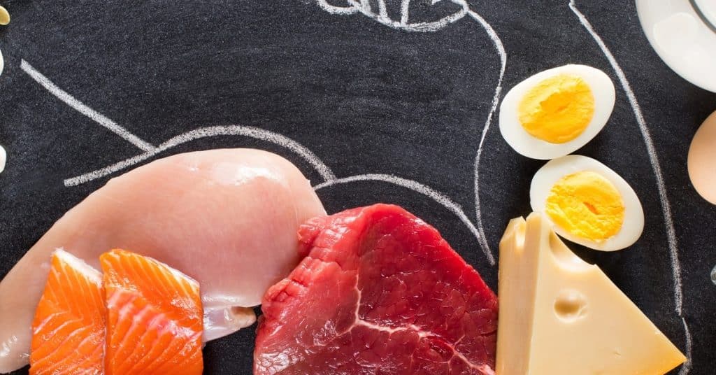 A black chalkboard features a drawing of a flexed bicep surrounded by foods like salmon, chicken, eggs, and cheese.