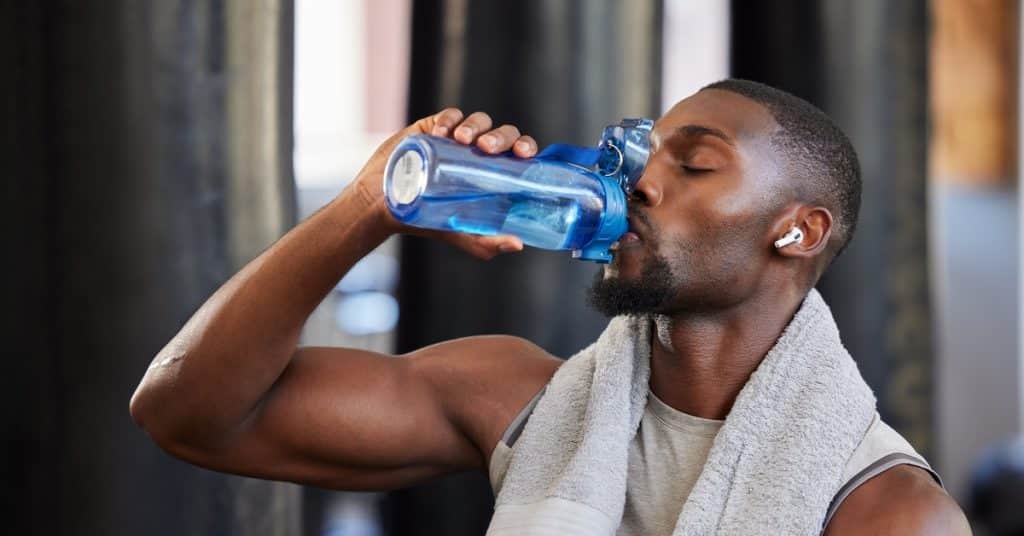 A young Black man drinks water from a blue bottle. He has headphones on and a gray towel around his shoulders.