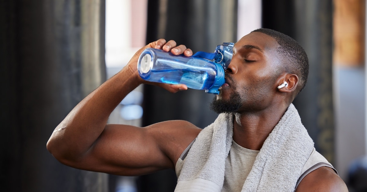 A young Black man drinks water from a blue bottle. He has headphones on and a gray towel around his shoulders.