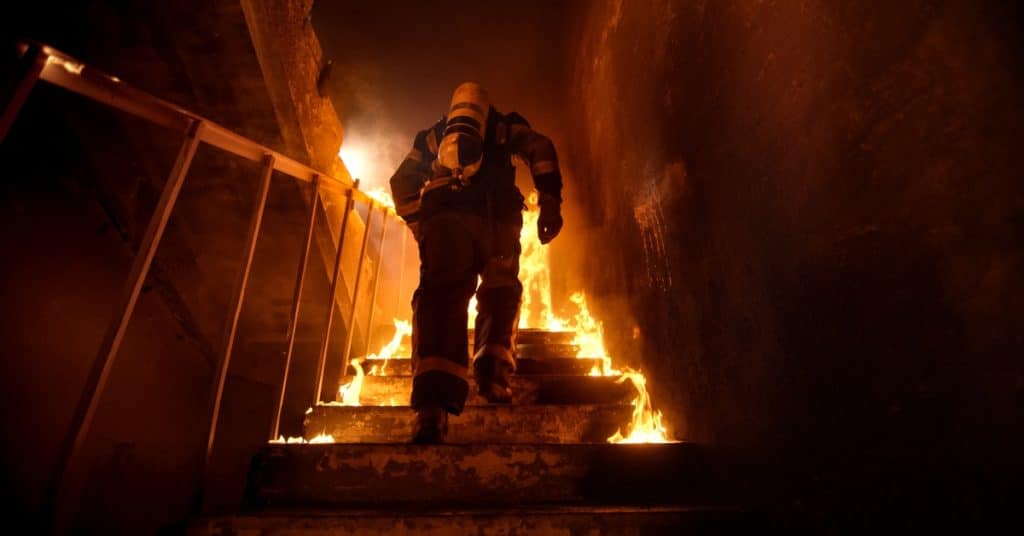 The back of a firefighter wearing a suit walking up stairs inside a darkly lit building that's surrounded by fire.