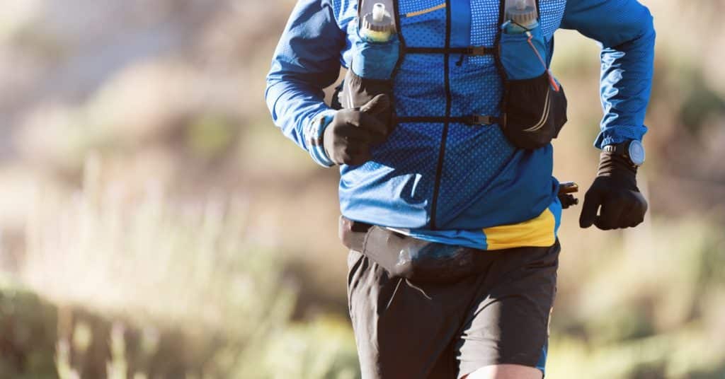 A man in workout clothes and a blue jacket runs through a remote area with a hydration backpack and black gloves.